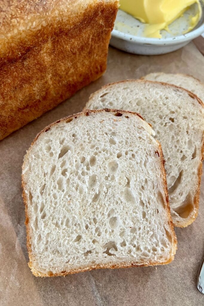 Sourdough baked in a loaf pan and sliced up. There is a butter dish in the background of the photo.