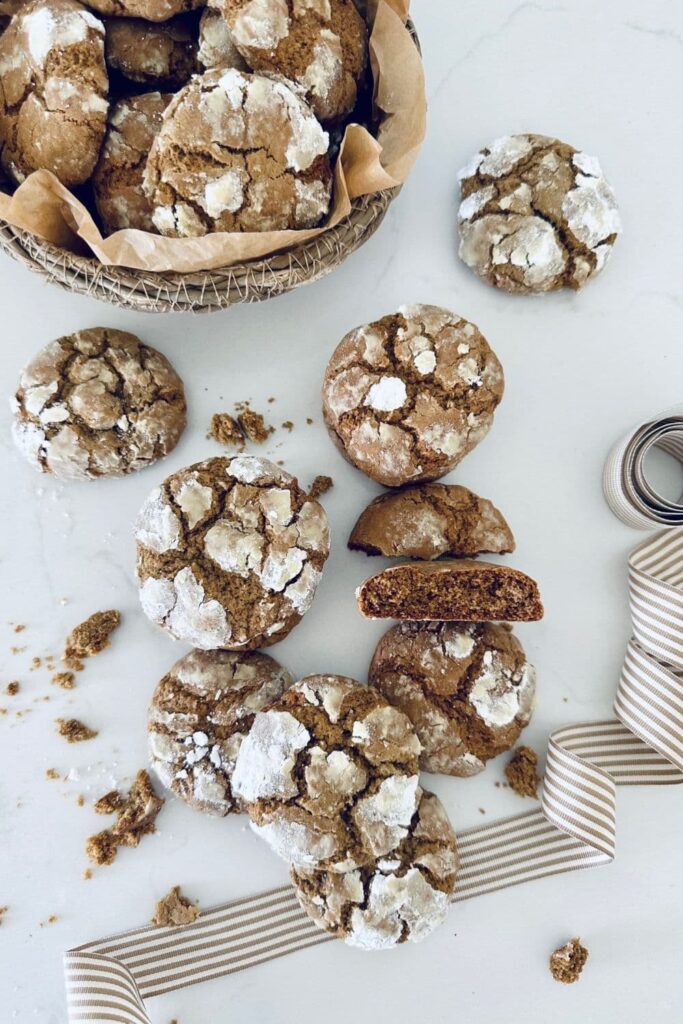 A pile of sourdough gingerbread crinkle cookies sitting on a white benchtop. You can see a basket of cookies at the top of the photo.