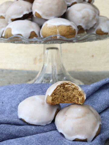 Sourdough pfeffernusse cookies sitting on a glass cake plate. There is a blue dish towel underneath with a few pfeffernusse sitting on them.