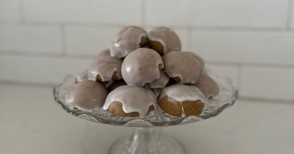 A pile of sourdough pfeffernusse cookies sitting on a glass cake plate in front of a white tiled wall.