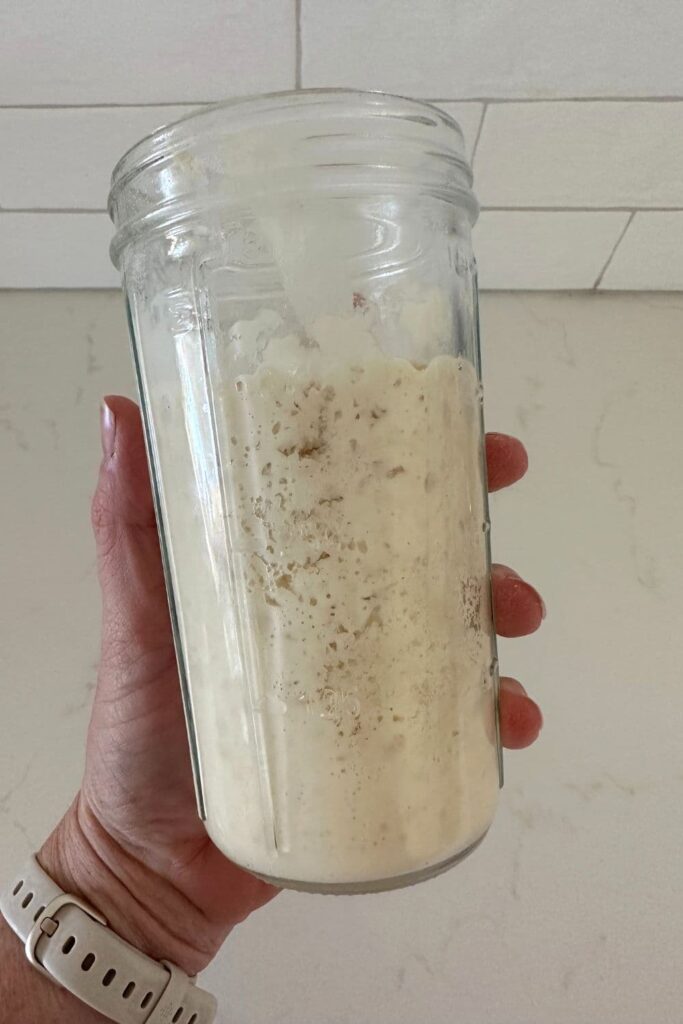 A jar of sweet stiff sourdough levain being held by a hand up against a white tiled wall.