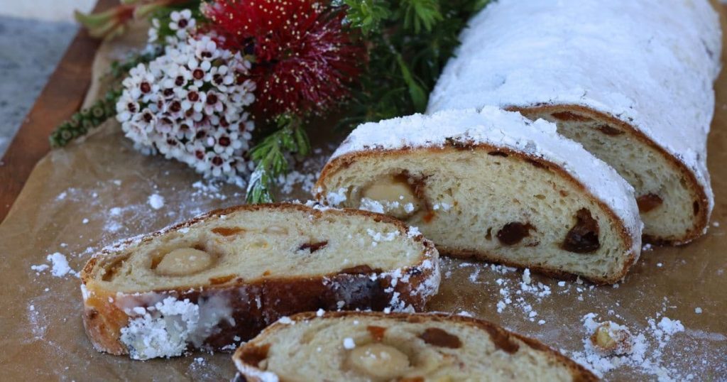 A traditional sourdough stollen dusted in white powedered sugar. The first few slices of the sourdough stollen have been sliced up to show the marzipan and dried fruit inside. There is a bunch of red and white Christmas flowers to the right of the sourdough stollen.