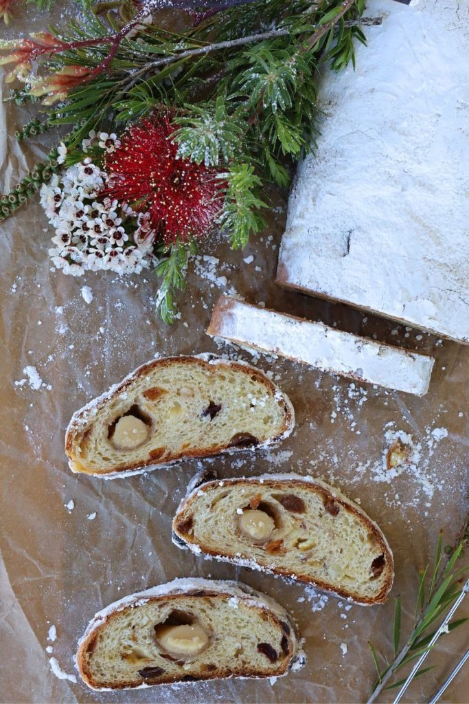 A traditional sourdough stollen dusted in white powedered sugar. The first few slices of the sourdough stollen have been sliced up to show the marzipan and dried fruit inside. There is a bunch of red and white Christmas flowers to the right of the sourdough stollen.