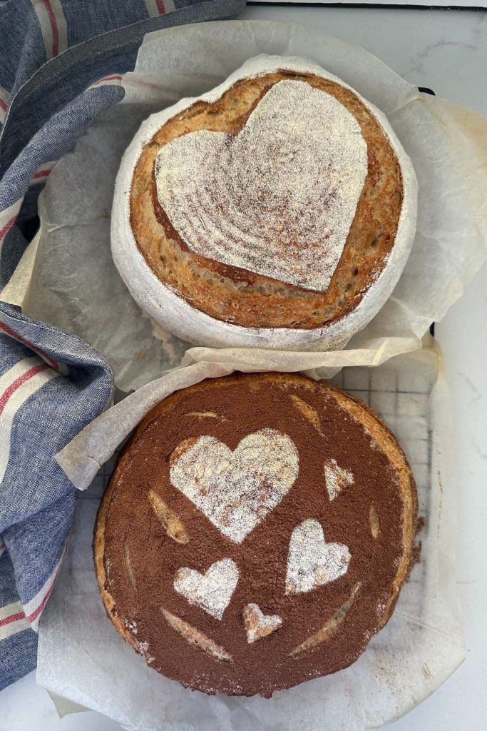 Two loaves of sourdough bread for Valentine's Day. The top one has a white heart scored into it and the bottom one has white hearts stencilled on surrounded by cocoa powder.