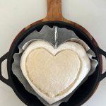 Heart shaped sourdough bread sitting in a Lodge Cast Iron Dutch Oven skillet lid. The dough has been scored with a large heart and has some aluminium foil in the Dutch Oven to help the bread heart retain its shape.
