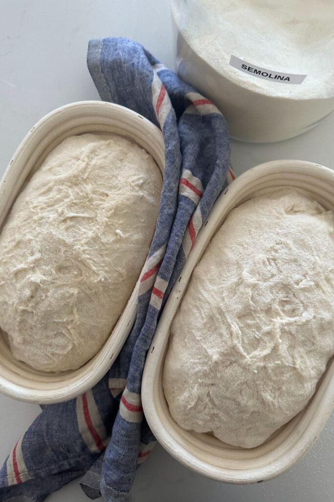 2 loaves of sourdough sitting in banneton baskets. There is a jar of semolina flour in the background.