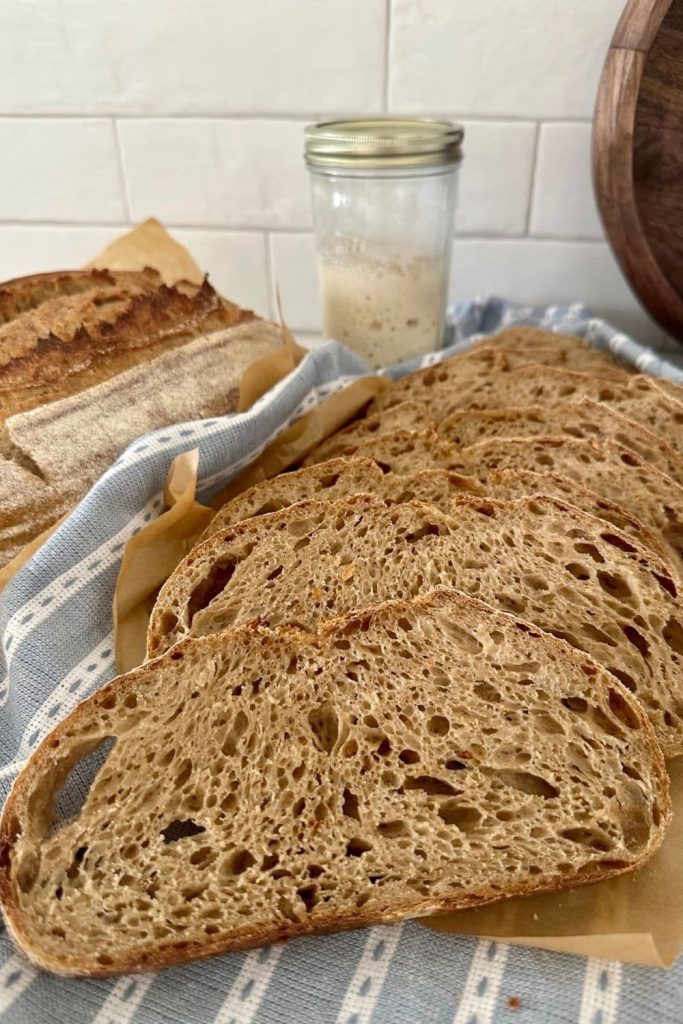 Sourdough country loaf sliced into slices. There is a jar of sourdough starter and another whole loaf of sourdough country loaf in the background.