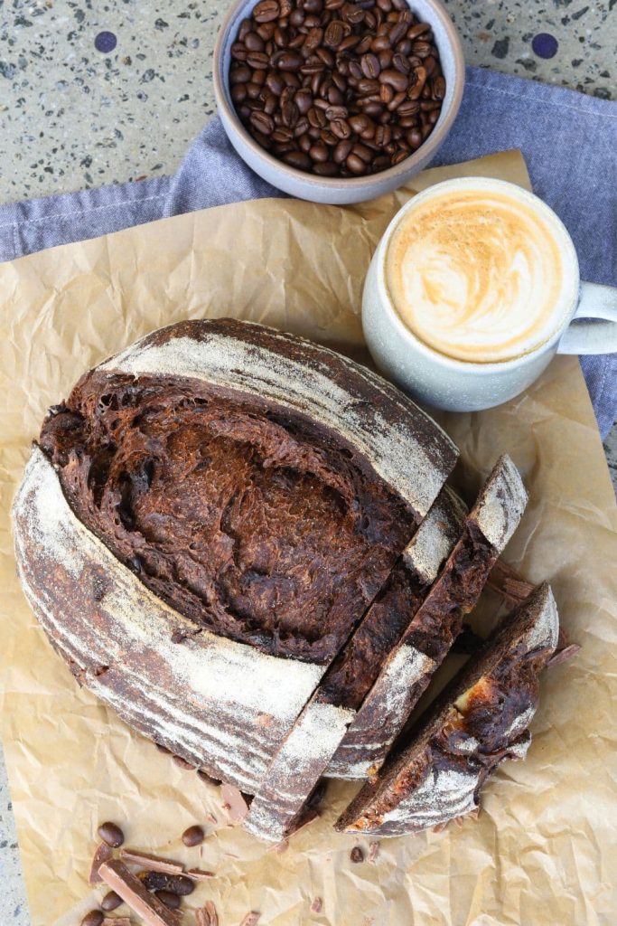A chocolate sourdough loaf sitting on a sheet of brown parchment paper next to a cup of coffee and bowl of coffee beans.