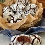 Sourdough chocolate crinkle cookies displayed in a basket lined with parchment paper. You can see a green plant in the background, as well as a blue dish towel.