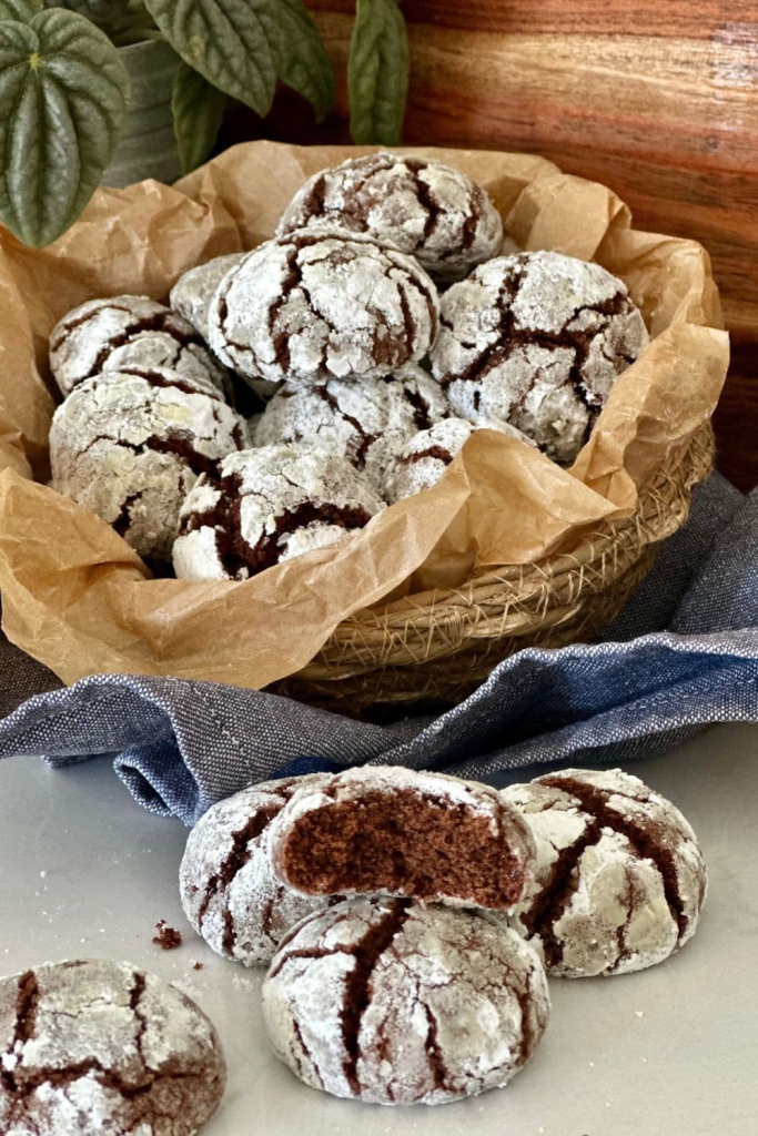 Sourdough chocolate crinkle cookies displayed in a basket lined with parchment paper. You can see a green plant in the background, as well as a blue dish towel.