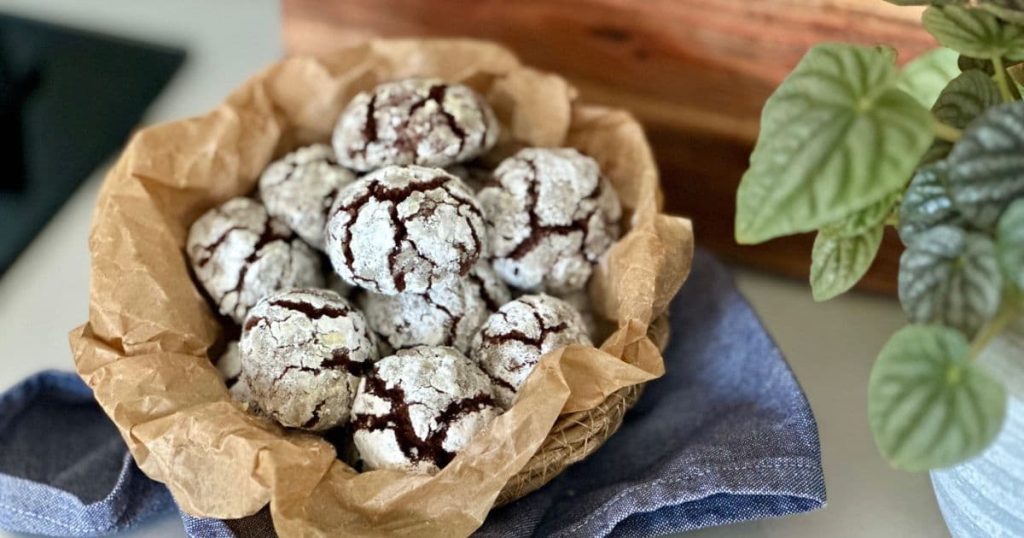 Sourdough chocolate crinkle cookies displayed in a basket lined with parchment paper. You can see a green plant in the background, as well as a blue dish towel.