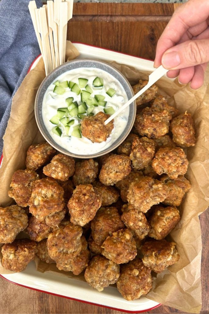 A tray of sourdough sausage balls presented with a small bowl of tzatziki. The sourdough sausage balls are being eaten with a wooden skewer.