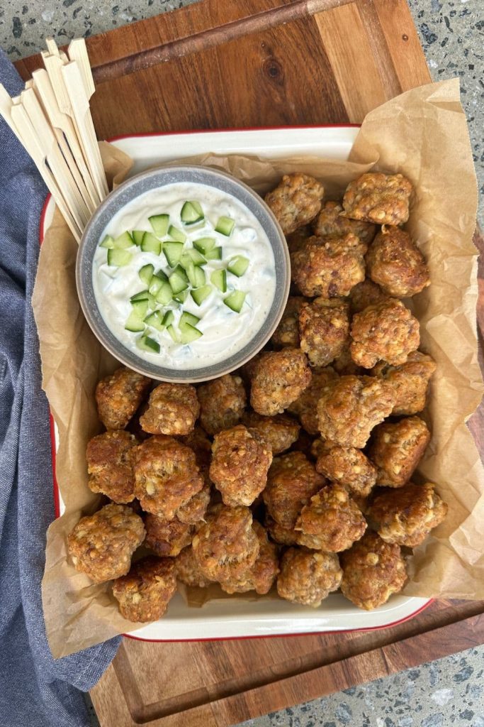 A tray of sourdough sausage balls presented with a small bowl of tzatziki. The sourdough sausage balls are being eaten with a wooden skewer.