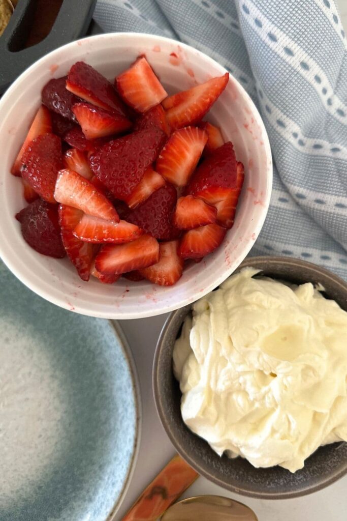 A bowl of strawberries and a bowl of vanilla whipped cream. There is a blue striped dishtowel to the right of the bowls.