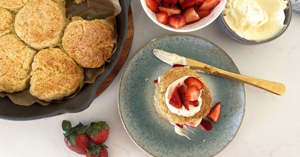 A cast iron skillet with sourdough biscuits baked inside. There is a sourdough strawberry shortcake assembled in the foreground, with bowls of sugared strawberries and whipped cream in the background.