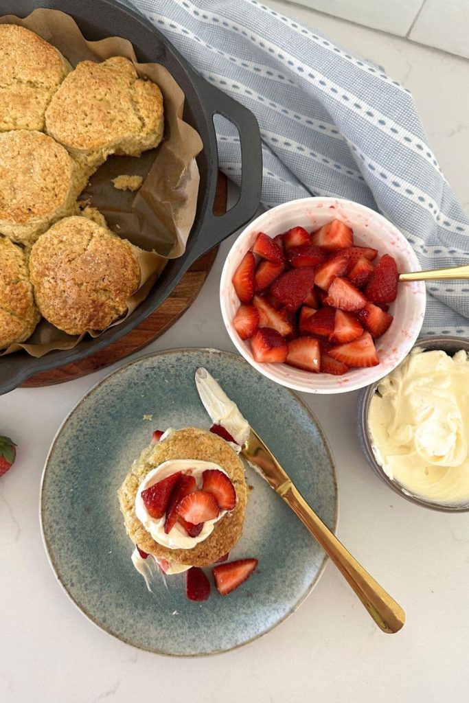 A cast iron skillet with sourdough biscuits baked inside. There is a sourdough strawberry shortcake assembled in the foreground, with bowls of sugared strawberries and whipped cream in the background.