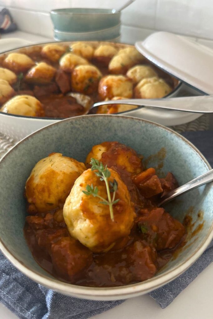 A blue bowl filled with sourdough Irish stew and topped with sourdough herb dumplings. You can see a large pot of sourdough irish stew and dumplings in the background of the photo.