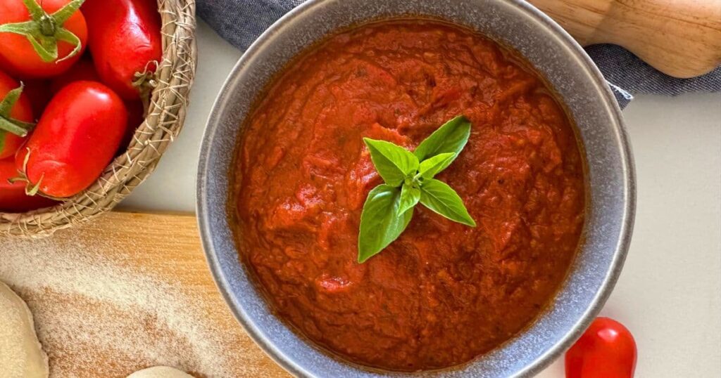 Bowl of homemade pizza sauce ready to be spread on sourdough pizza. There are a few small basil leaves in the centre of the homemade pizza sauce and you can see some fresh tomatoes in a basket to the left of the sauce.