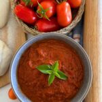 Homemade pizza sauce made using fresh roma tomatoes. The pizza sauce is displayed in a grey stoneware bowl with a piece of green basil in the centre. You can also see some pizza dough, a bowl of roma tomatoes and a rolling pin in the photo.