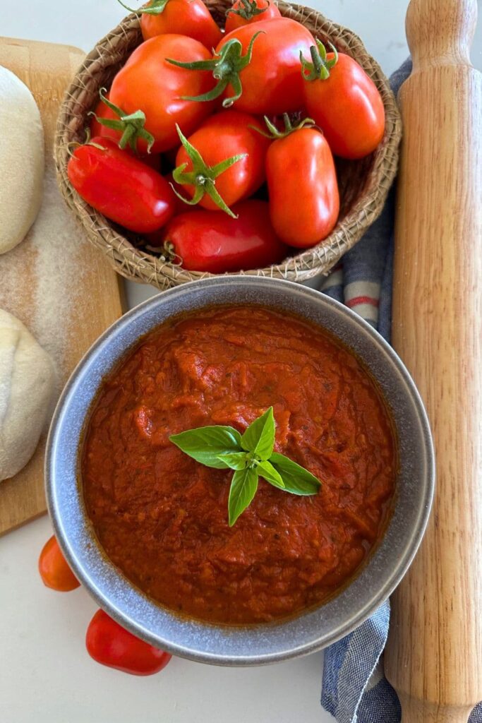 Homemade pizza sauce made using fresh roma tomatoes. The pizza sauce is displayed in a grey stoneware bowl with a piece of green basil in the centre. You can also see some pizza dough, a bowl of roma tomatoes and a rolling pin in the photo.