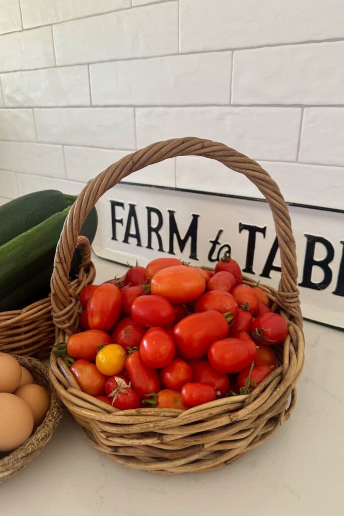 A cane basket filled with red roma tomatoes. You can see a sign that says "Farm to Table" in the background, as well as a basket of zucchinis and eggs.