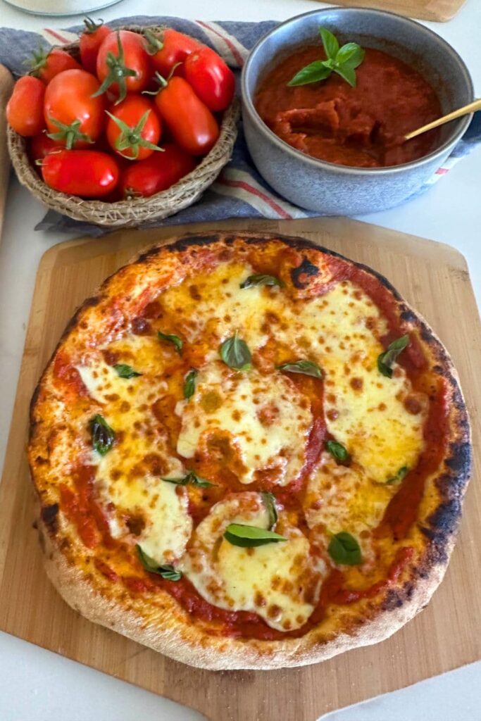 A sourdough pizza that has been topped with homemade pizza sauce displayed on a wooden board. You can also see a bowl of roma tomatoes and a bowl of pizza sauce towards the top of the photo.