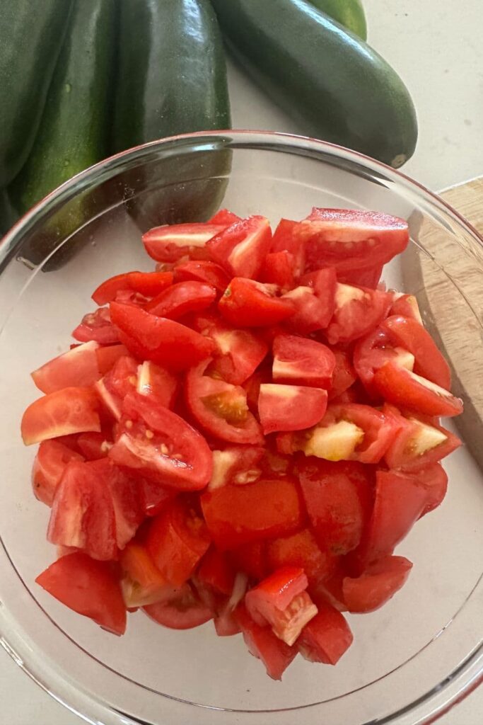 A glass bowl of diced Roma Tomatoes. You can see some whole green zucchini in the background.