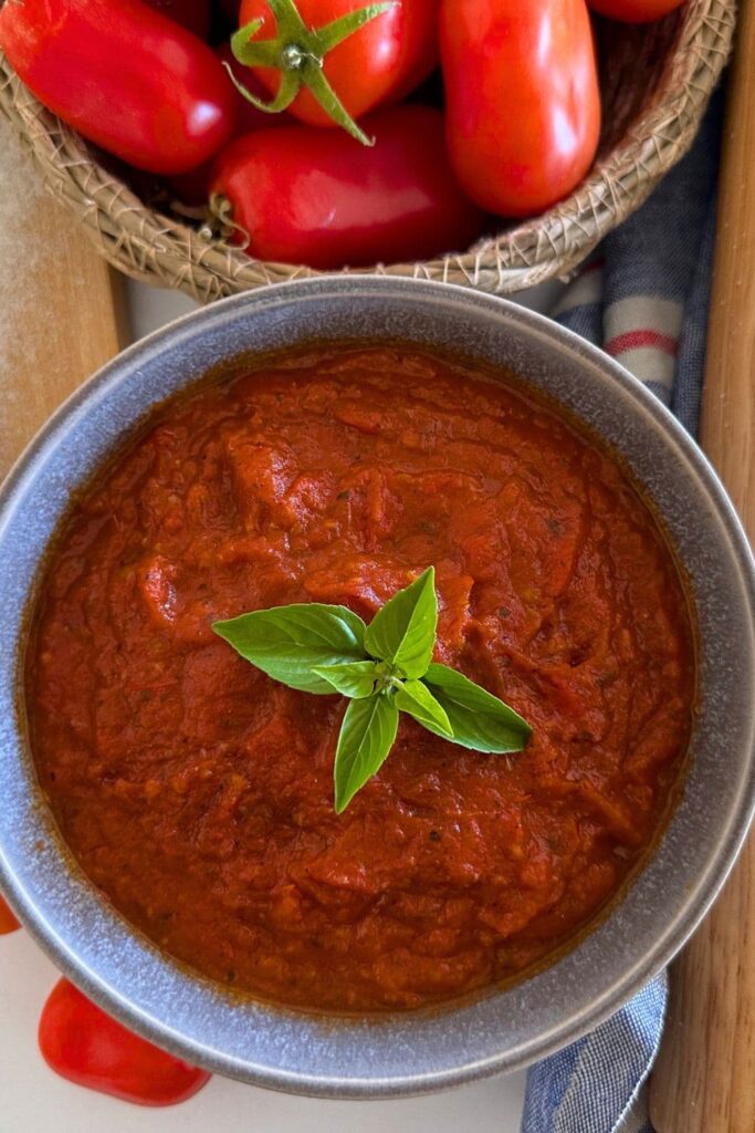 A grey stoneware bowl filled with homemade pizza sauce. There is a piece of green basil in the centre of the sauce for decoration. You can see a bowl of red roma tomatoes at the top of the photos.