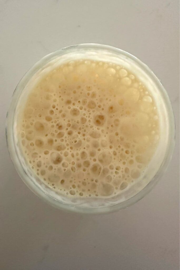 A photo of a jar of sourdough starter taken from above the jar so you can see the top of the starter. The top of the sourdough starter has foamy, soap like bubbles and is nearly reaching the rim of the jar.