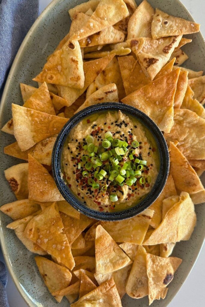 A bowl of hummus made without tahini sitting in the centre of a green platter and surrounded by sourdough pita chips. The hummus has been topped with chopped green shallots, chili flakes and olive oil. 