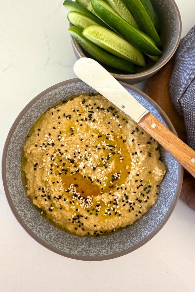 A bowl of hummus made without tahini and topped with sesame seeds and nigella seeds. There is a spead knife sitting on the top of the bowl. You can see some cucumber sticks in the background.