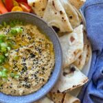 A bowl of hummus made without tahini. It's served in the middle of a platter surrounded by sourdough flat bread, red and yellow tomatoes and is topped with chopped shallots.