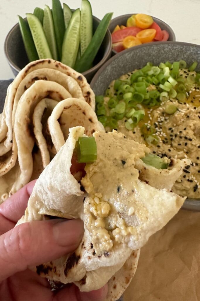 A sourdough flat bread that has been scrunched up and dipped into the bowl of hummus which is sitting behind. You can also see some colorful vegetable sticks in the background of the photo.