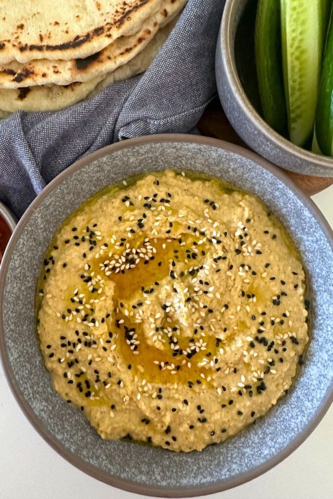 A grey stoneware bowl filled with hummus made without tahini. The hummus is topped with nigella seeds and sesame seeds. You can see some sourdough flat bread and cucumber sticks in the background.