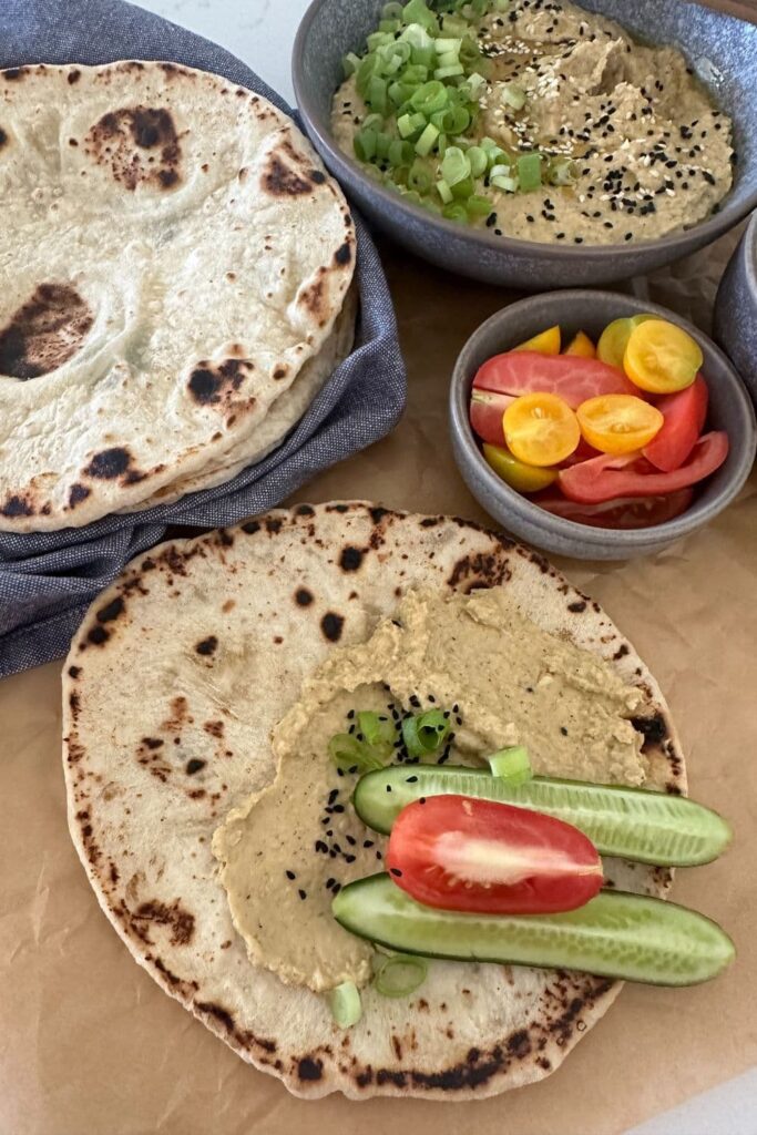A sourdough flat bread topped with some creamy hummus, cucumber sticks and tomatoes. You can see a stack of sourdough flat breads in the background, along with a bowl of hummus made without tahini.
