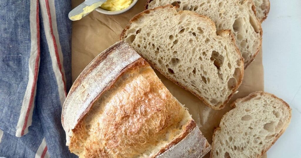 A loaf of extra sour sourdough bread that has had a few slices cut from it. It is displayed on a piece of parchment paper beside a blue striped linen dish towel.