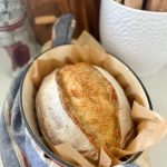 A loaf of extra sour sourdough bread nestled in parchment paper inside a cream enamel Dutch Oven. There is a blue striped linen dish towel threaded through the handles of the Dutch Oven and big pot of rolling pins in the background of the photo also.