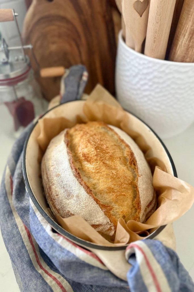 A loaf of extra sour sourdough bread nestled in parchment paper inside a cream enamel Dutch Oven. There is a blue striped linen dish towel threaded through the handles of the Dutch Oven and big pot of rolling pins in the background of the photo also.