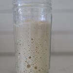 A jar of sourdough starter sitting on a white counter top. The jar has large bubbles on the side of the glass.