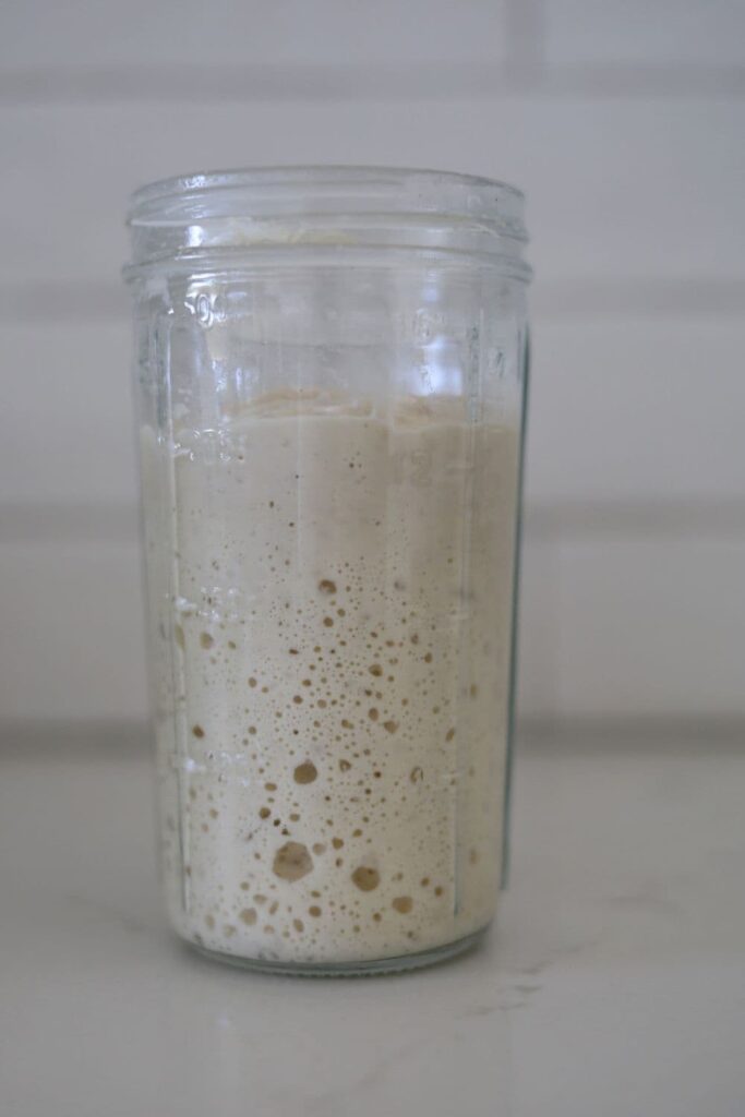 A jar of sourdough starter sitting on a white counter top. The jar has large bubbles on the side of the glass.