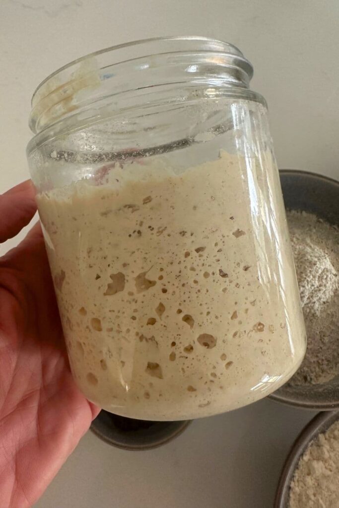 A jar of sourdough starter being held up to the light so you can see the bigger sourdough bubbles on the side of the jar.