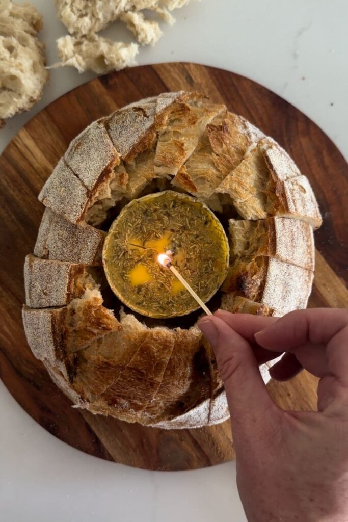 A butter candle sitting in the middle of a loaf of sourdough bread. You can see a hand lighting the candle in the centre of the bread with a small match.