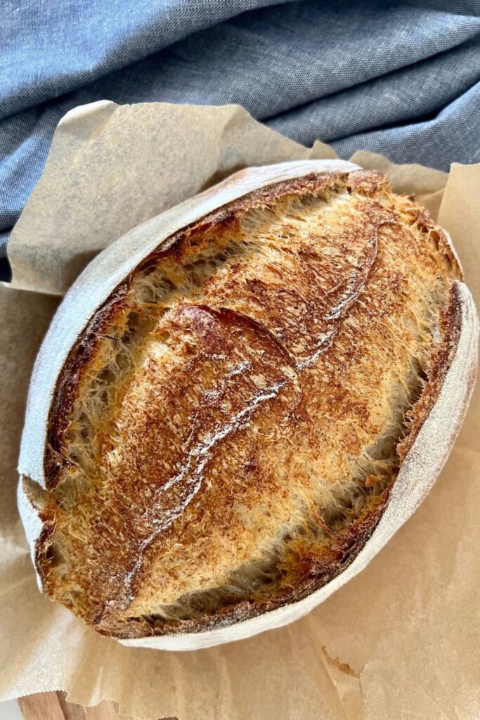 A large loaf of sourdough bread sitting on a piece of parchment paper. There is a blue dish towel in the background.