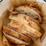 A loaf of sourdough bread that has been slice and is nestled in a piece of parchment paper inside a cream enamel Dutch Oven.