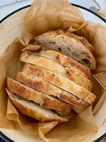 A loaf of sourdough bread that has been slice and is nestled in a piece of parchment paper inside a cream enamel Dutch Oven.