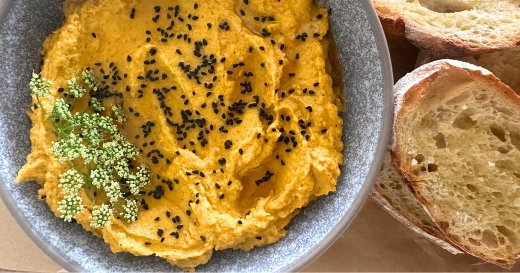 ROASTED VEGETABLE HUMMUS SERVED IN A GREY STONEWARE BOWL NEXT TO SOME SOURDOUGH CROSTINI.