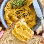 A bowl of roasted vegetable hummus decorated with cumin seeds. The bowl is surrounded by sourdough crostini and there is a slice of sourdough crostini spread with roasted carrot hummus in the foreground.