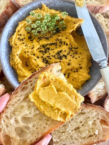 A bowl of roasted vegetable hummus decorated with cumin seeds. The bowl is surrounded by sourdough crostini and there is a slice of sourdough crostini spread with roasted carrot hummus in the foreground.