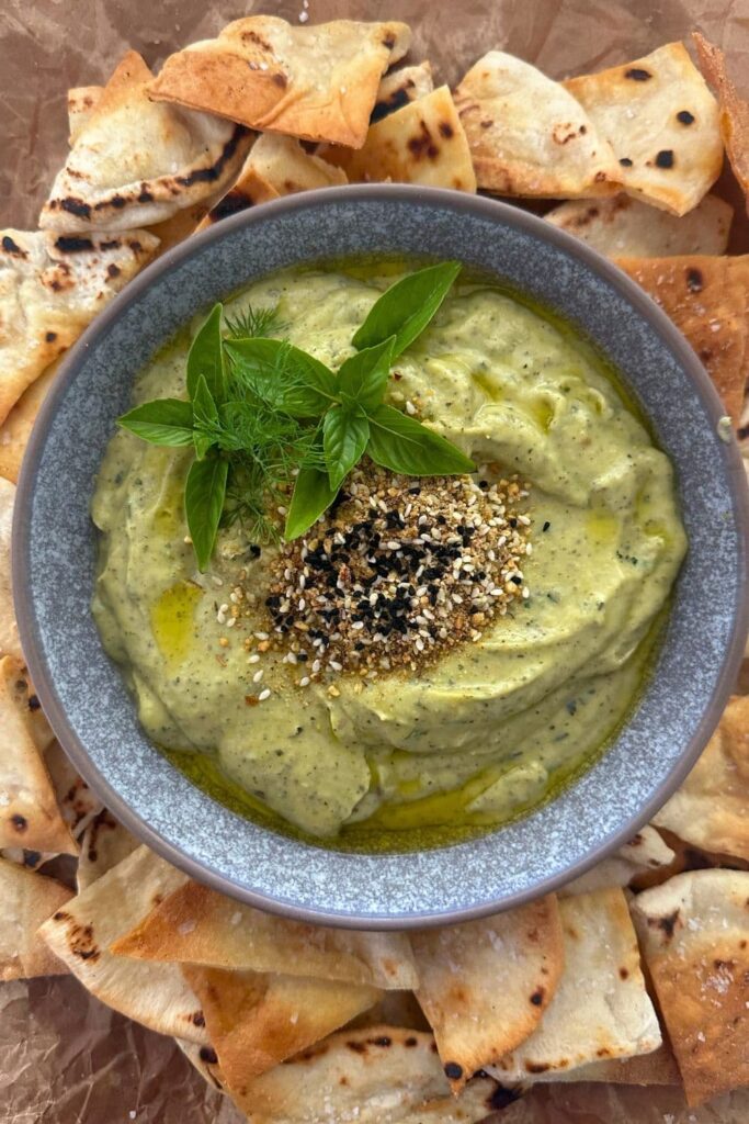 Zucchini yogurt dip topped with dukkah, olive oil and fresh herbs and served in a grey stoneware bowl. The bowl of zucchini yogurt dip is surrounded by sourdough pita chips.