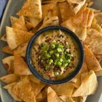 Sourdough Pita Chips displayed on a green glazed dish. There is a bowl of hummus in the centre that has been topped with shallots and cumin seeds.